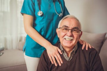 Imagebild zum Pflegebonus - älterer Herr sitzt, hinter ihm steht eine Krankenschwester und hält beruhigend ihre Hände auf seine Schultern