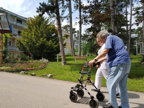 2 Menschen gehen spazieren. Einer davon ist älter und benutzt einen Rollator